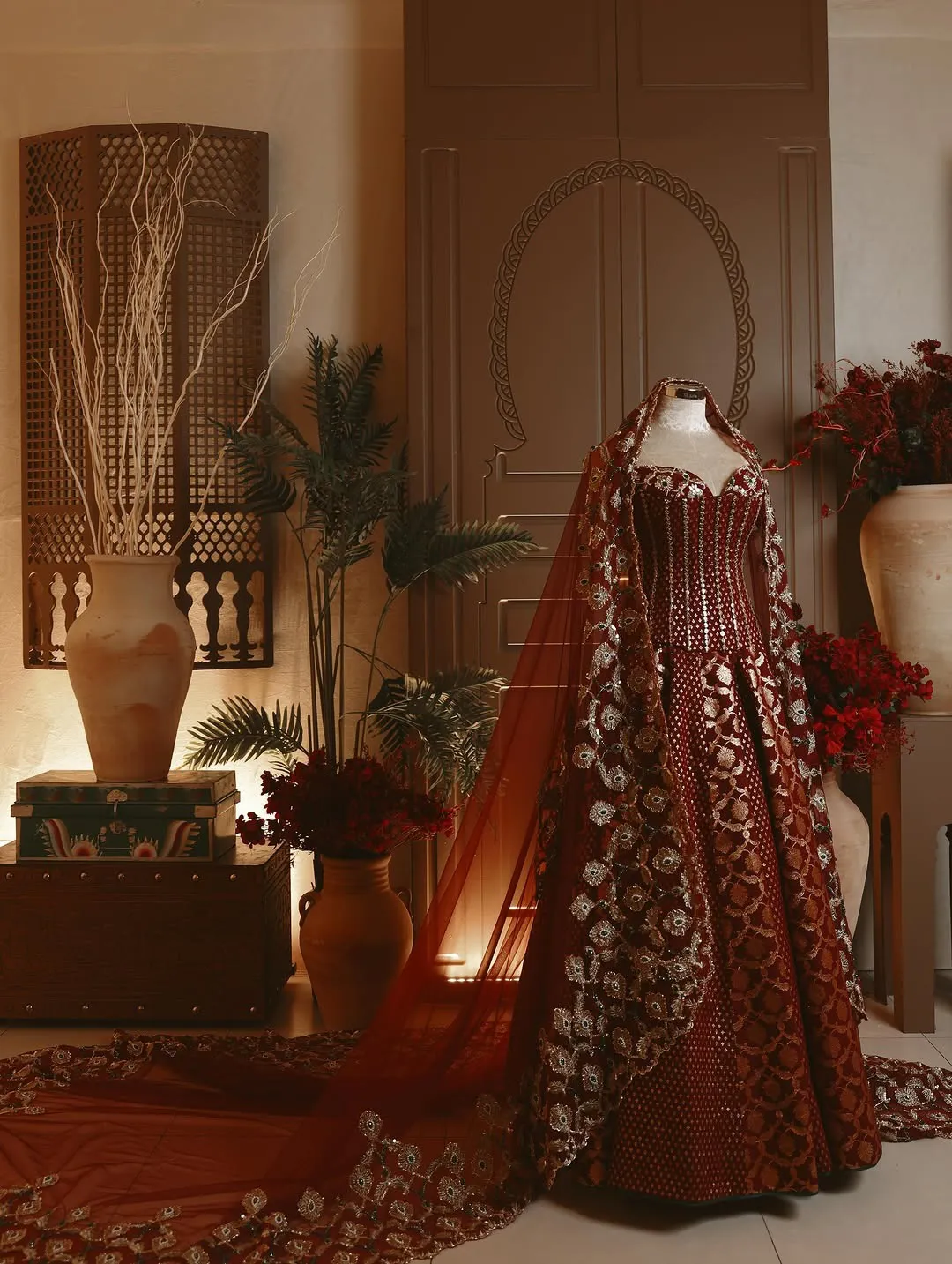 A red and gold bridal gown with a long embroidered veil, set among clay jars, palms and a carved door