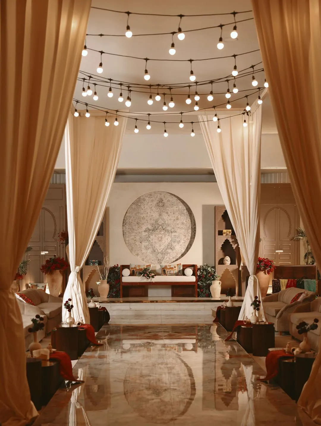 A draped entrance under string lights opening onto a majlis daybed, clay jars and a polished marble aisle